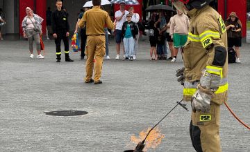Strażak podpalający garnek z paliwem. W tle budynek straży oraz zebrani ludzie.